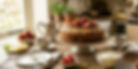 Cake topped with strawberries and nuts on a rustic table, set with tea, cream, and lemons. Cozy kitchen in the background.
