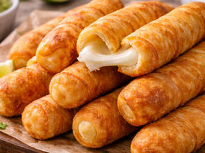 Golden, crispy cheese sticks stacked on parchment paper, one oozing melted cheese. Lime wedge and green sauce in the background.