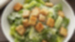 Bowl of Caesar salad with romaine, croutons, and grated cheese. Rustic wooden table, creamy dressing visible, cozy dining vibe.