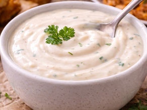A white bowl of creamy dip garnished with parsley, surrounded by fried plantains and appetizers on a wooden table.