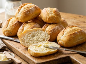 Fresh bread rolls piled on a wooden board, one split open with butter, on a rustic table beside cups and a butter dish.