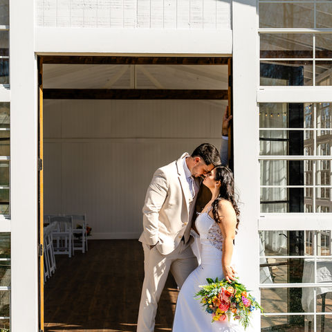 Newlywed couple sharing an intimate embrace at Violets Venue- person in cream suit gently holding person in white lace wedding dress with colorful bouquet in the door way.