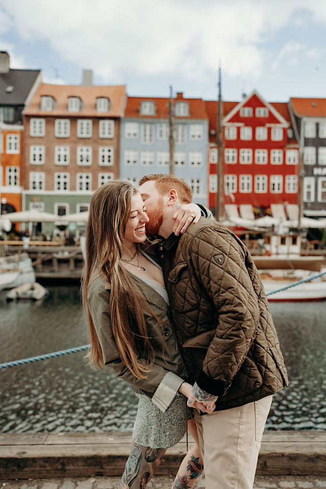Newly engaged couple walking hand in hand along the Nyhavn waterfront in Copenhagen, smiling at each other with the full row of colourful historic canal houses behind them