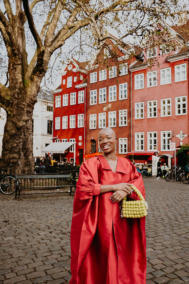 Sofiyat Ibrahim standing under a tree in Gråbrødretorv, sunlight filtering through the leaves, highlighting her red dress.