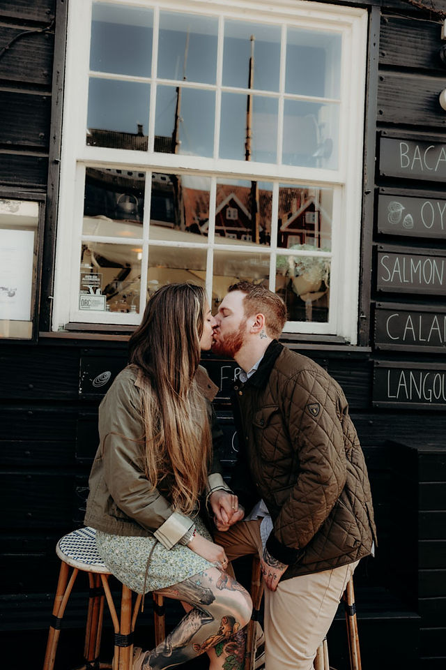 Newly engaged couple walking hand in hand along the Nyhavn waterfront in Copenhagen, smiling at each other with the full row of colourful historic canal houses behind them