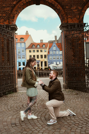 Newly engaged couple walking hand in hand along the Nyhavn waterfront in Copenhagen, smiling at each other with the full row of colourful historic canal houses behind them