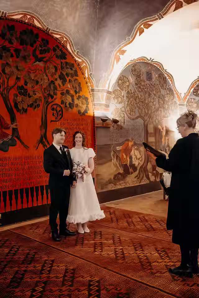 Bride and groom standing together during their Copenhagen City Hall ceremony as the officiant reads, framed by vibrant mural paintings.