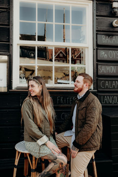 Newly engaged couple walking hand in hand along the Nyhavn waterfront in Copenhagen, smiling at each other with the full row of colourful historic canal houses behind them