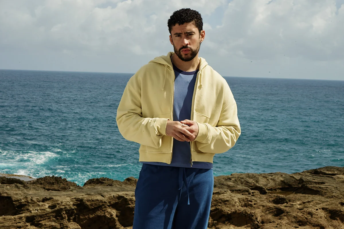 “Un verano en NUEVAYoL”… con Benito aterrizando en España. Coincidiendo con el arranque de sus conciertos en España, la marca de Inditex lanza BENITO ANTONIO, una cápsula de casi 150 piezas que traduce el universo estético del artista puertorriqueño a prendas pensadas para el día a día. Oversize, sastrería relajada, camisetas gráficas, bermudas amplias, bandanas y tejidos ligeros forman parte de una propuesta que bebe directamente de Puerto Rico y de esa manera despreocupada —pero muy medida— de vestir que define a Benito. Lejos de parecer el típico merchandising de gira, la colección funciona como una extensión real de su estilo personal: colores vibrantes, siluetas cómodas y ese punto entre urbano y nostálgico que domina también sus últimos trabajos musicales. Además, llega en uno de los momentos de mayor locura, con fans agotando prendas en cuestión de horas y preparando looks para los conciertos de Madrid y Barcelona.