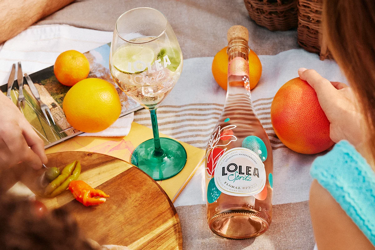 LOLEA    Lolea entra en este universo con una propuesta que apuesta por sabores mediterráneos, ingredientes naturales y una estética tan cuidada como la propia bebida. Su gama Lolea Spritz nace alrededor de dos versiones muy distintas, pero igual de apetecibles: Citrus Spritz y Floral Spritz. La primera combina vino blanco espumoso con notas de bergamota, cítricos y un toque aromático de albahaca que la hace especialmente fresca y ligera. La segunda se mueve en un terreno más suave y floral, mezclando vino rosado, naranja sanguina y azahar en una combinación delicada y ligeramente ácida que funciona especialmente bien al atardecer. Ambas referencias tienen una graduación moderada del 8% y están elaboradas mediante el método Charmat, responsable de esa burbuja fina y elegante que hace que cada sorbo resulte más suave.  