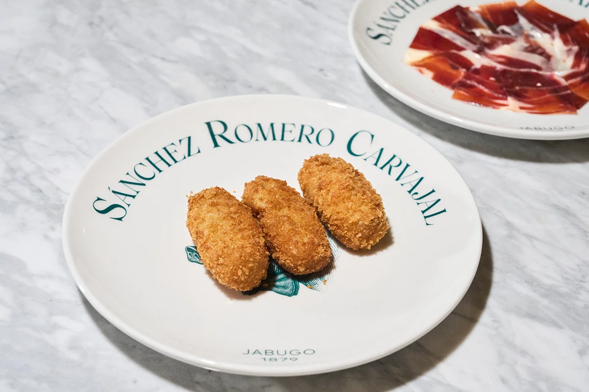 Ruta de la Croqueta de Sánchez Romero Carvajal La croqueta es uno de esos clásicos siempre presente en los mejores momentos de tapeo y terraceo. Los amantes de esta elaboración están de enhorabuena porque ya pueden disfrutar de la Ruta de la Croqueta de Sánchez Romero Carvajal, celebración que cumple su cuarta edición con una propuesta más amplia y con más fuerza que nunca. Hasta el 6 de julio, este recorrido se instala como una de las citas gastronómicas más esperadas del inicio del verano con los nuevos participantes como Salino, Los Gabrieles, Árdia, Rita Sibarita, Casa Vega, Cachivache (Serrano), La Raquetista, Cachivache (Montecarmelo) y El Gallego, que amplían el itinerario con nuevas interpretaciones de este clásico. Las incorporaciones se suman a algunos de los grandes nombres de otras ediciones como Casa Orellana, Tatel, Casa Salesas, Café Comercial, 11 Nudos, El Escondite de Villanueva o Virrey. Además, con motivo del Día Mundial de la Tapa, la ruta amplía su propuesta con una irresistible selección de tapas elaboradas con jamón ibérico.