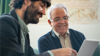 Two colleagues from different generations sit side by side, smiling and looking at a tablet, as the younger person points something out—capturing a moment of knowledge sharing or reverse mentoring in a workplace setting.