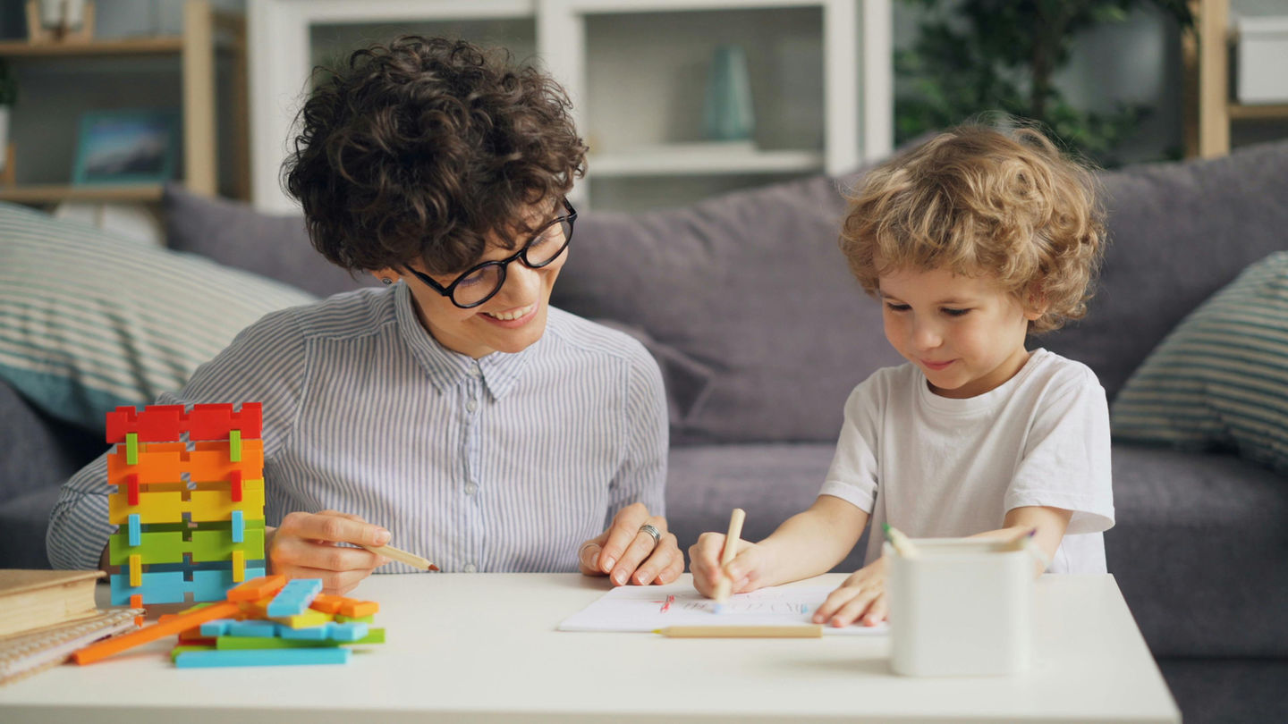 Service de garde d’enfants à domicile à Paris : femme souriante et enfant bouclé dessinant à table avec des blocs colorés.