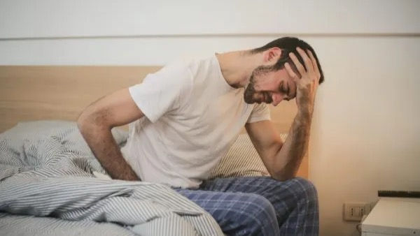 Man sitting on the side of his bed, holding his head in agony. He looks defeated as if he's in pain or dealing with stress.