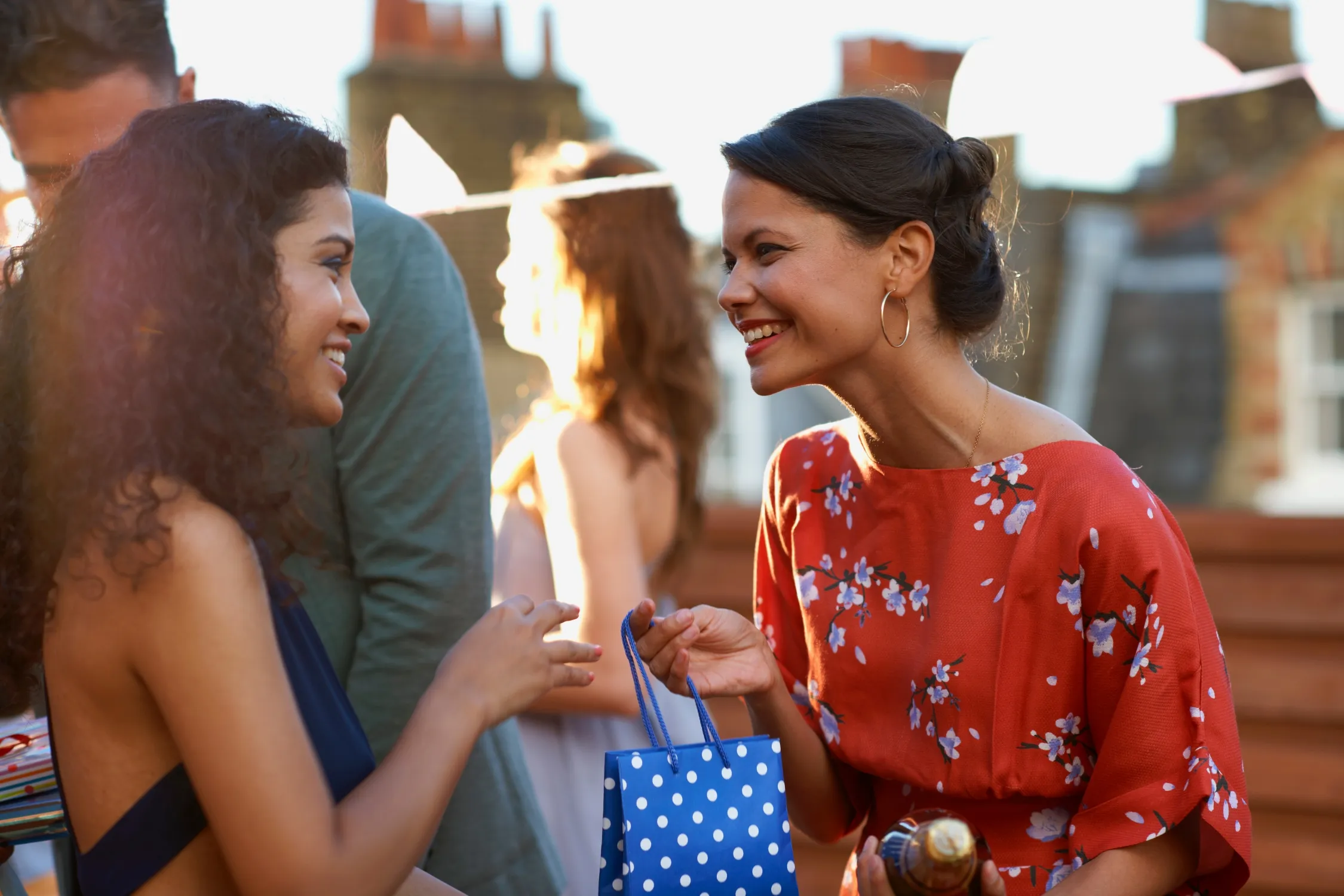 Woman ina red dress with white flowers giving a unique gift in a blue and white dot bag to another woman in a navy blue dress with big smiles as thankful for the best gift