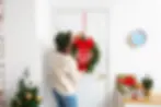 Una persona añade un toque festivo a su hogar colgando una exuberante corona navideña verde adornada con frutos rojos en una clásica puerta blanca. La acogedora escena navideña incluye un pequeño árbol decorado que brilla con luces, regalos cuidadosamente envueltos en diversos papeles alegres y un encantador cartel con la palabra "XMAS" colocado en una mesa cercana, lo que crea una atmósfera acogedora y alegre.