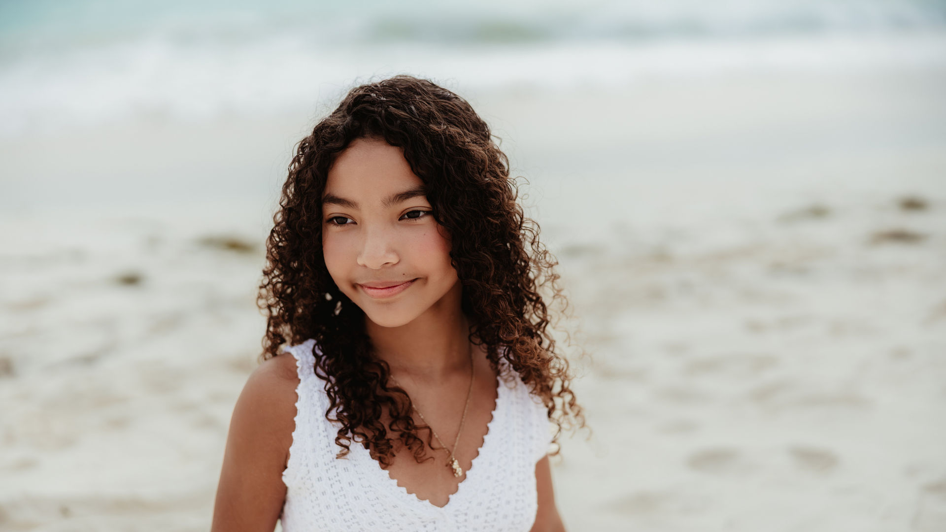 Girl with curly hair sitting in the white sand on the beach in Aruba, wearing a white dress during a natural outdoor photoshoot