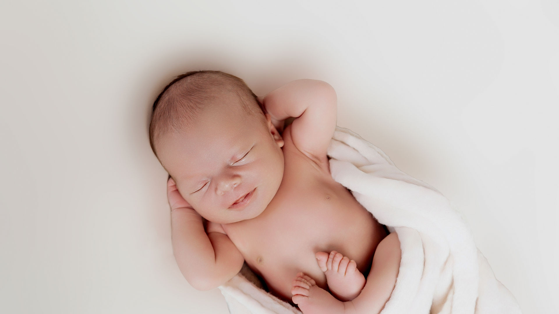 Smiling newborn baby boy with hands behind his head during an indoor studio photoshoot in Aruba, wrapped on a bean bag in neutral tones