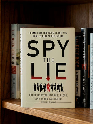A paperback copy of the book "Spy the Lie" by Philip Houston, Michael Floyd, and Susan Carnicero, displaying its beige cover with the subtitle "Former CIA Officers Teach You How to Detect Deception." It is standing upright on a wooden bookshelf next to other books, creating a realistic study or library setting.
