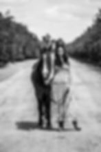 A teenage girl posing with her horse in black and white. She's wearing a dress while standing on a dirt road with horse at her side.
