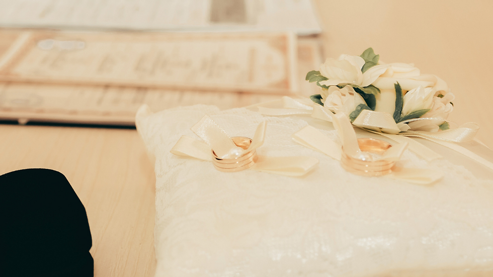Gold wedding rings on a lace pillow with ribbon, a floral corsage, and a blurred background of certificates on a light wooden table.
