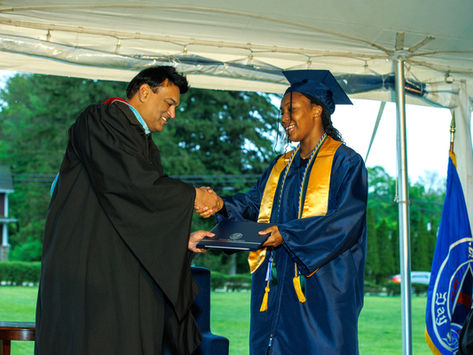 Saddle River Day School graduate in blue gown and cap accepts diploma with a handshake outdoors, surrounded by trees and a blue flag. Celebratory mood.