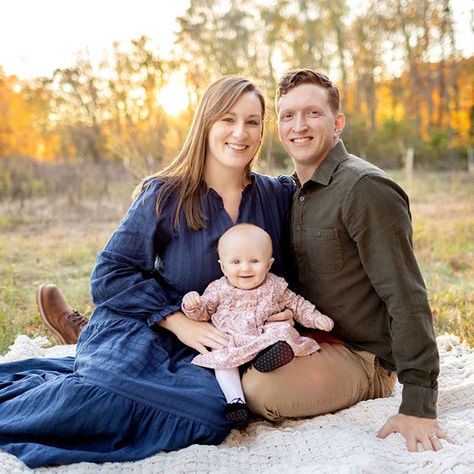 Smiling parents sit on a blanket with their baby in a sunlit autumn field, with trees glowing gold in the background.