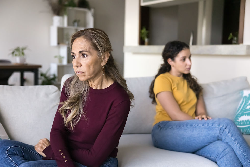 Two women sit back-to-back on a couch in a bright living room, looking tense and upset, in maroon and yellow tops.