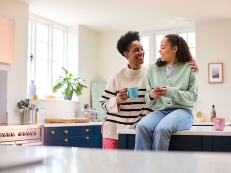 Two smiling women in a bright kitchen, one holding a blue mug, the other a phone. Plant on windowsill, cozy and cheerful atmosphere.