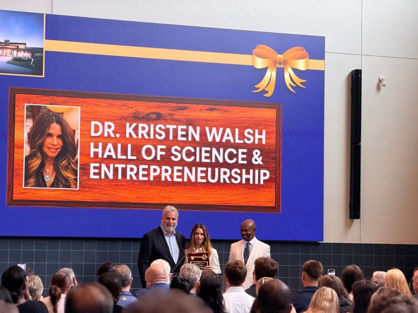 Woman holds plaque on stage with two men at Dr. Kristen Walsh Hall of Science & Entrepreneurship ceremony before crowd.