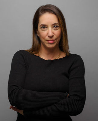 Portrait of a woman in a black top with arms crossed, standing against a plain gray background with a calm, serious expression
