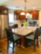 Kitchen with wooden cabinets, granite island, and black chairs. Yellow vase with flowers on the island. Silver appliances and hanging lights.