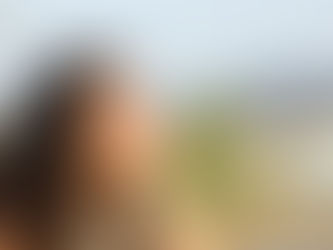 Smiling woman with curly hair reclines outdoors, eyes closed, enjoying the sun. Blurred natural background creates a peaceful mood.