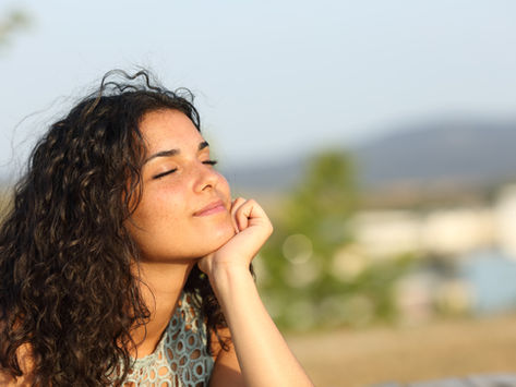 Smiling woman with curly hair reclines outdoors, eyes closed, enjoying the sun. Blurred natural background creates a peaceful mood.
