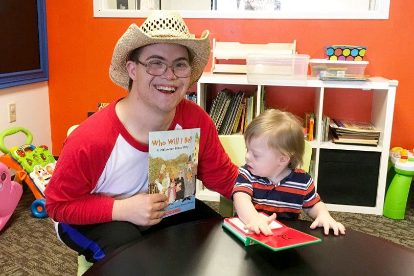Happy man in a cowboy hat holds Who Will I Be? beside a toddler at a table in a bright classroom.