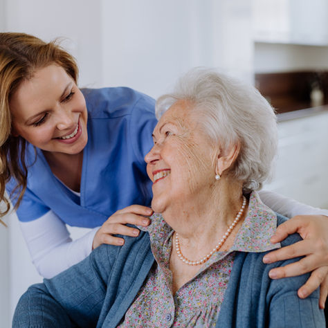 A caregiver in blue scrubs smiles warmly at an elderly woman in a wheelchair, inside a bright, white kitchen. They appear happy and engaged.