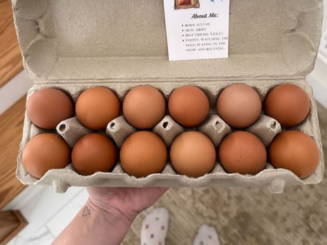 Hand holds a carton of brown eggs with a note about "Ruby." Background shows wooden stairs and person wearing white socks with dots.
