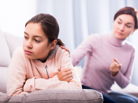 Upset girl sits on a couch while a woman comforts her in a bright living room, both in soft pink tops.