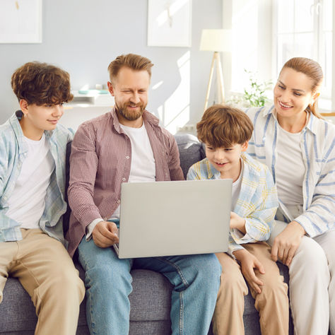 A family of four sits on a gray couch, smiling at a laptop. Bright, cozy living room with shelves and plants in the background.