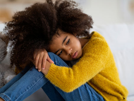 Child in yellow sweater sits curled up on a couch, looking sad. A gray stuffed animal is beside them. Warm tones create a serene mood.