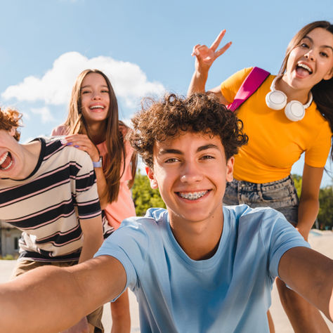 Four teens crowd a selfie outdoors under blue sky, smiling and flashing peace signs.
