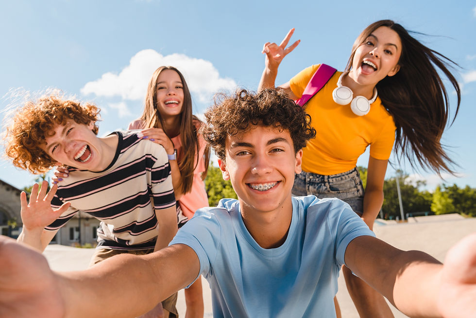 Four teens crowd a selfie outdoors under blue sky, smiling and flashing peace signs.