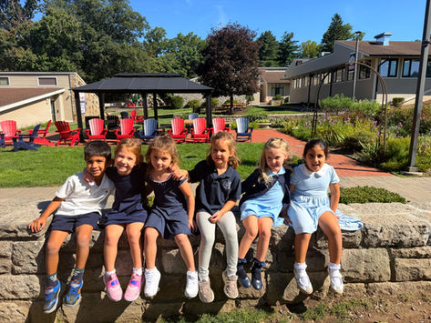 Six children sit smiling on a stone wall in a sunny school courtyard, with red chairs, gardens, and buildings behind.