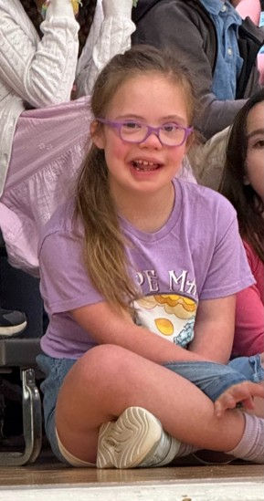 Young girl with glasses in a purple shirt and denim shorts, sitting cross-legged, smiling, in a crowd setting.