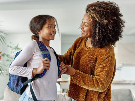 Smiling mother helps her daughter adjust a backpack in a bright home interior, both facing each other warmly.