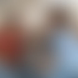 Two people sit on a gray couch in a kitchen, smiling at each other. One wears a red shirt, the other a blue shirt. Warm, relaxed mood.