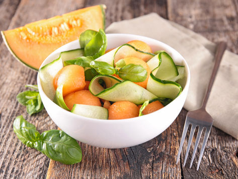 Cantaloupe and cucumber salad with basil in a white bowl on a wooden table. A fork and napkin are beside it. Fresh and vibrant setting.