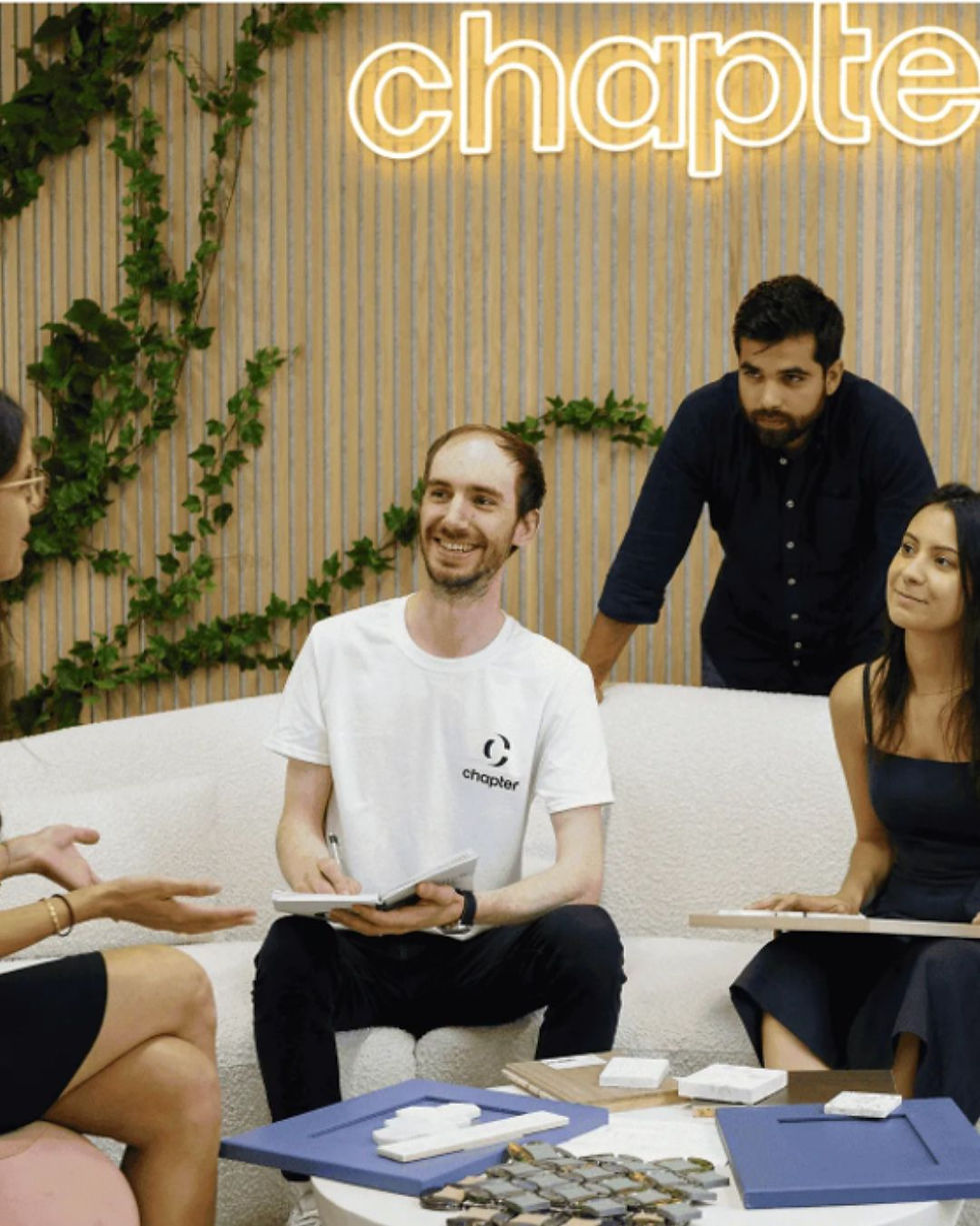People chat and take notes in a cozy lounge with a glowing chapter sign, plants, and sample cards on the table.