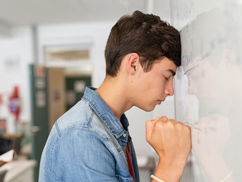 Teen in a denim jacket leans head against whiteboard covered in equations, eyes closed in frustration, in a classroom setting.