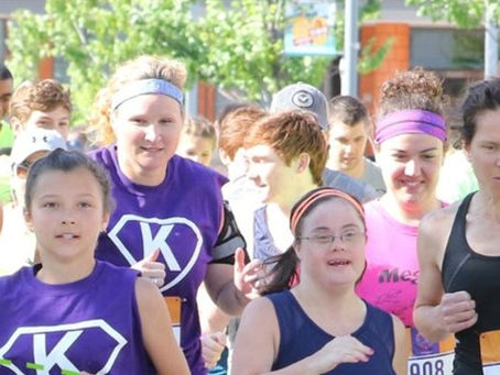 Group of runners race outdoors in bright sun, including women in purple K shirts and bibs, with focused expressions.